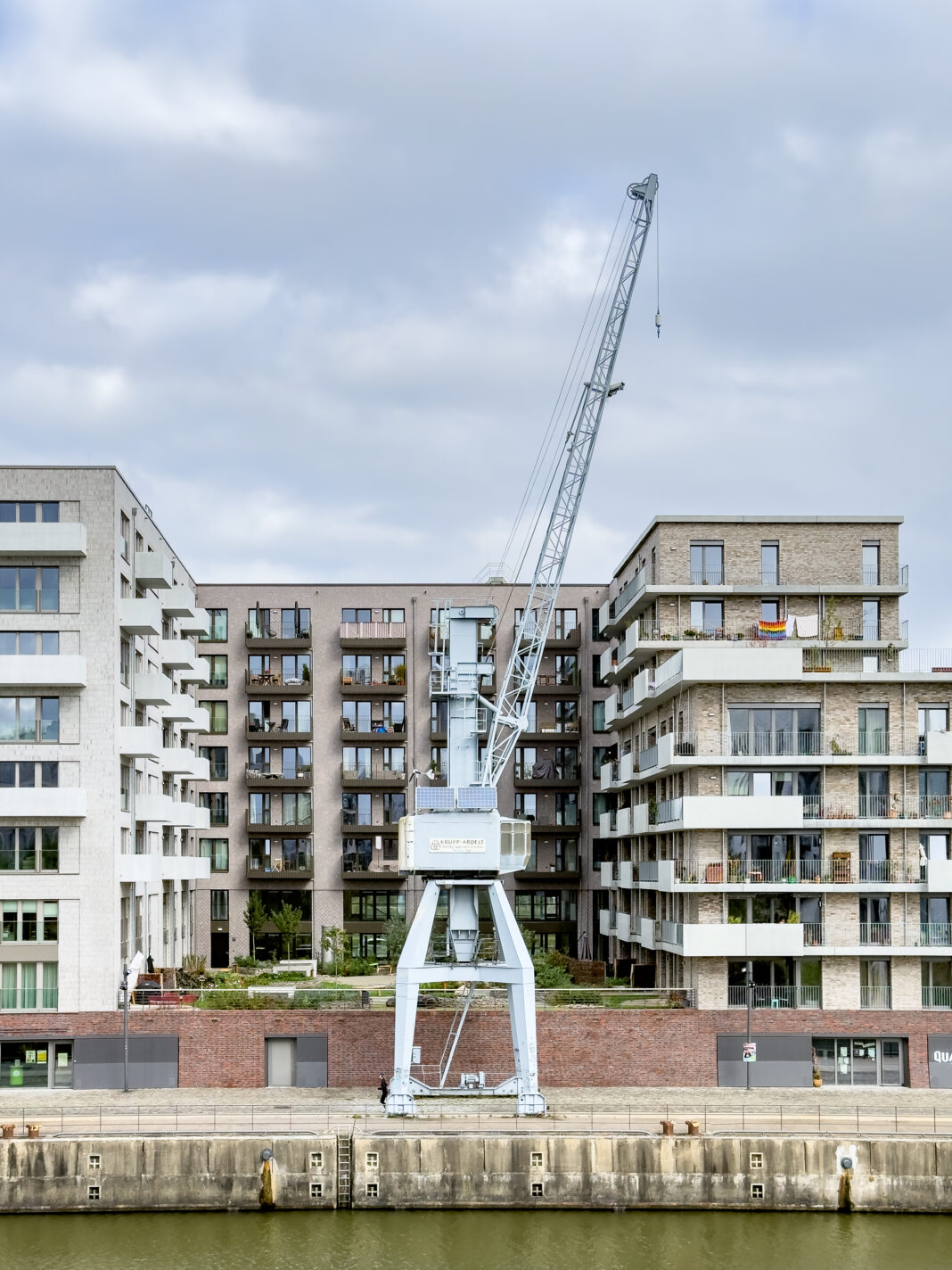 Foto: Kran in der Hafencity. Man sieht ein paar graue Neubauten vor grauem Himmel und mittig einen alten Kran. Der Kran ist auch grau.