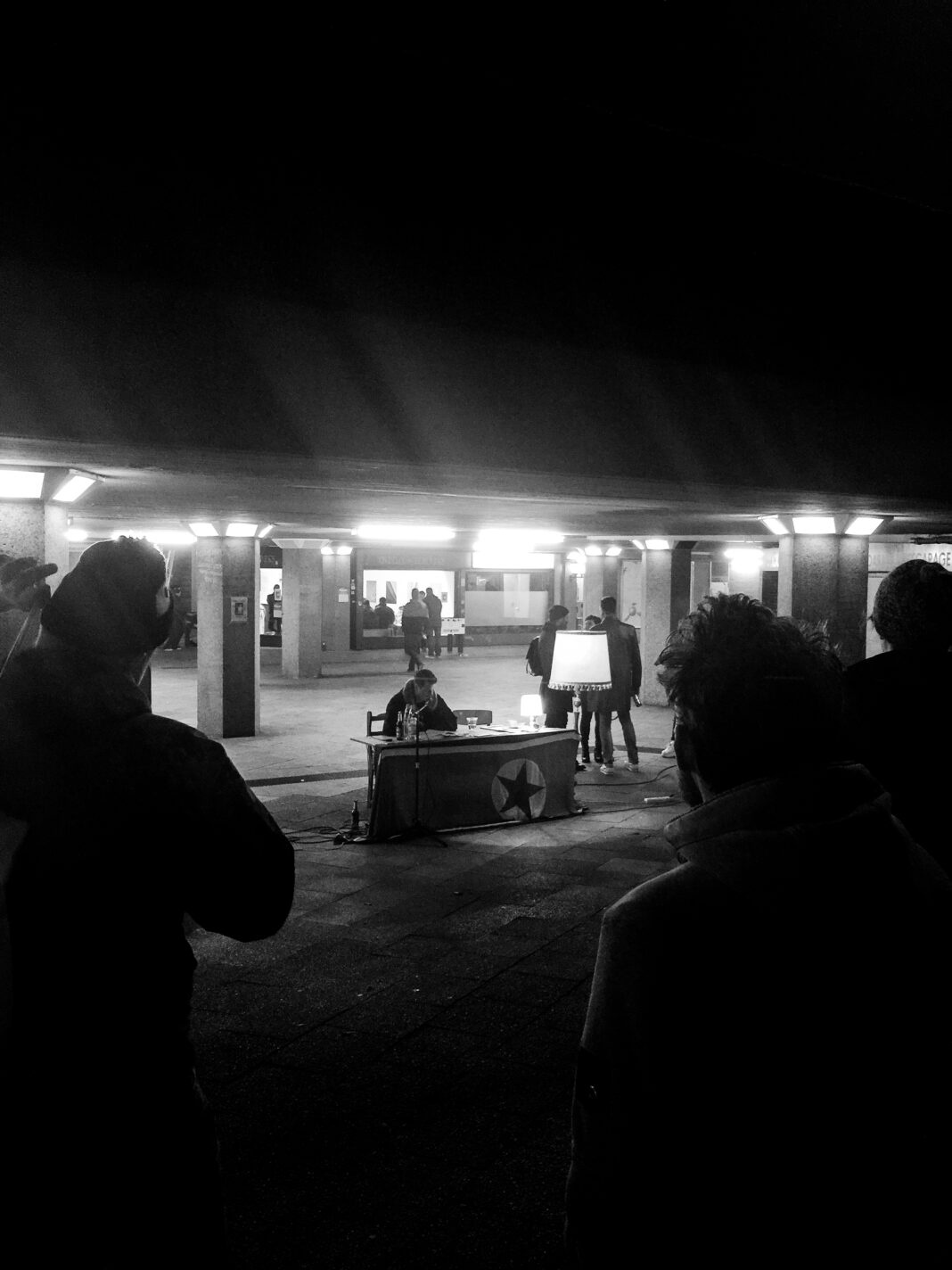 Eingerahmt von silhouettenhaft erscheinenden Zuhörern sitzt Sascha Bisley an einem Tisch am Ebertplatz, wo er an einem kalten Märzabend 2015 eine Lesung abhält.