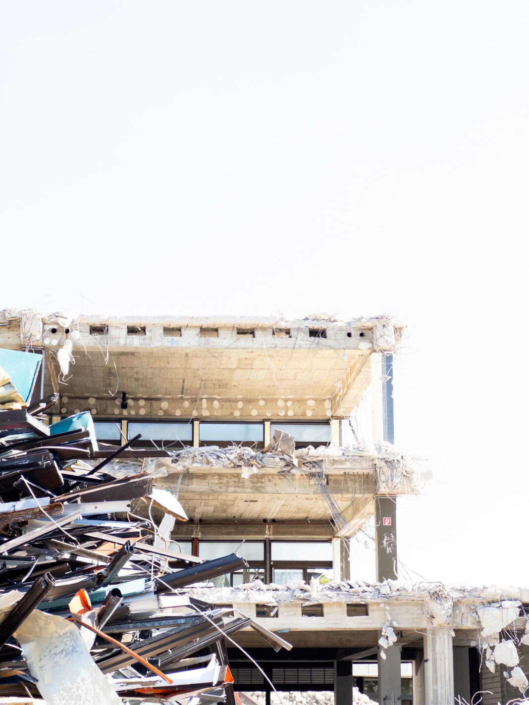 Auf einem überbelichteten sieht man vor einem farblosen Himmel das Gerippe des alten Schulkörpers, der von Baggern aufgerissen hinter Schutt und Schrott emporragt.