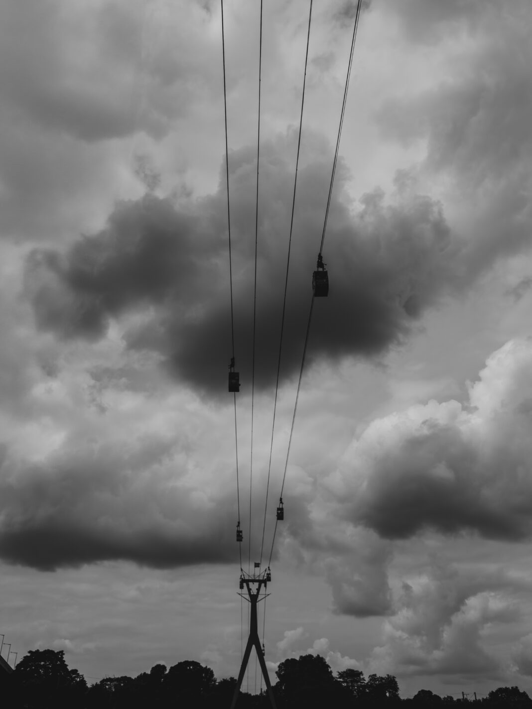 Foto: Kölner Seilbahn. Blick über den Rhein und unter den Seilen hindurch, an denen Gondeln hängen. Im HIntergrund ein wolkiger Himmel.