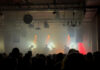 Foto: KMPFSPRT im Gebäude 9 Foto: KMPFSPRT im Gebäude 9. Blick auf die Bühne von weiter hinten im Saal. Alles recht diesig. Auf der Bühne steht die Band, im Saal freut sich das Publikum, dessen Hinterköpfe man erahnt.