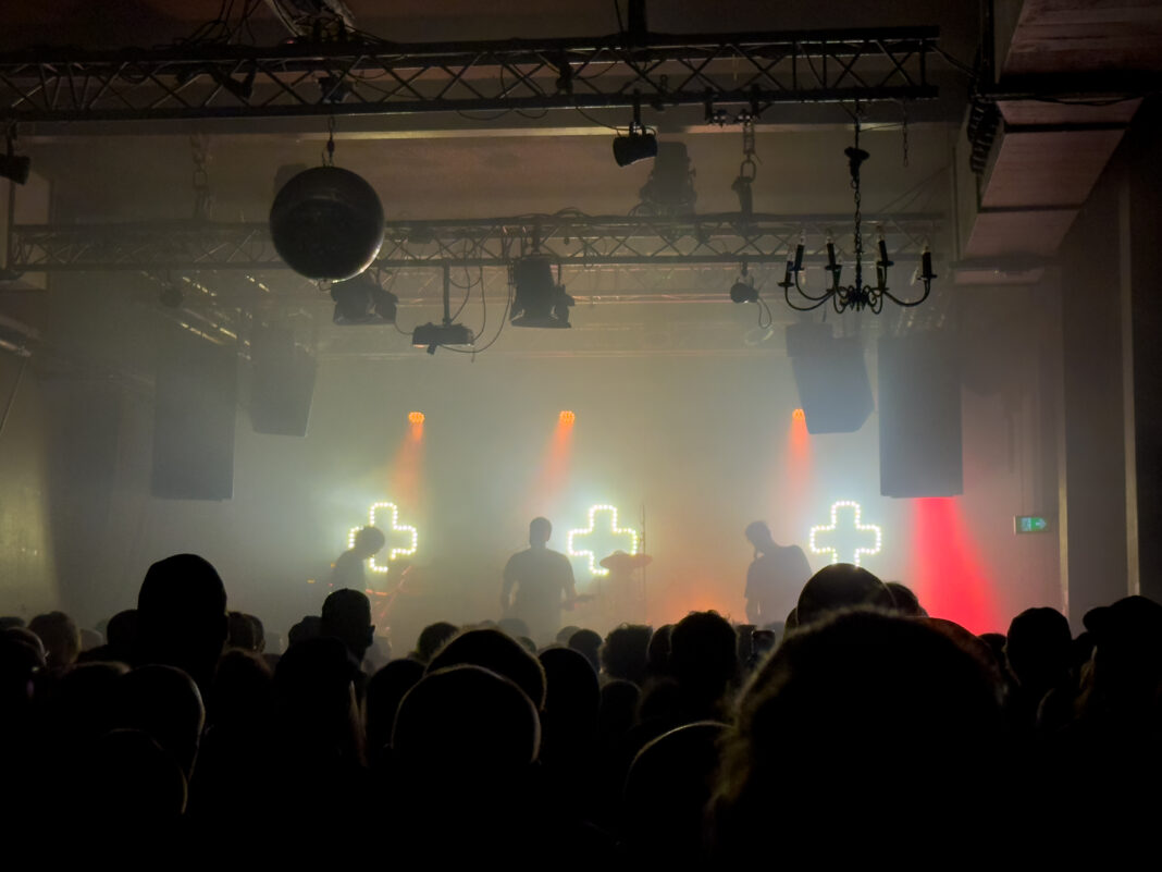 Foto: KMPFSPRT im Gebäude 9 Foto: KMPFSPRT im Gebäude 9. Blick auf die Bühne von weiter hinten im Saal. Alles recht diesig. Auf der Bühne steht die Band, im Saal freut sich das Publikum, dessen Hinterköpfe man erahnt.