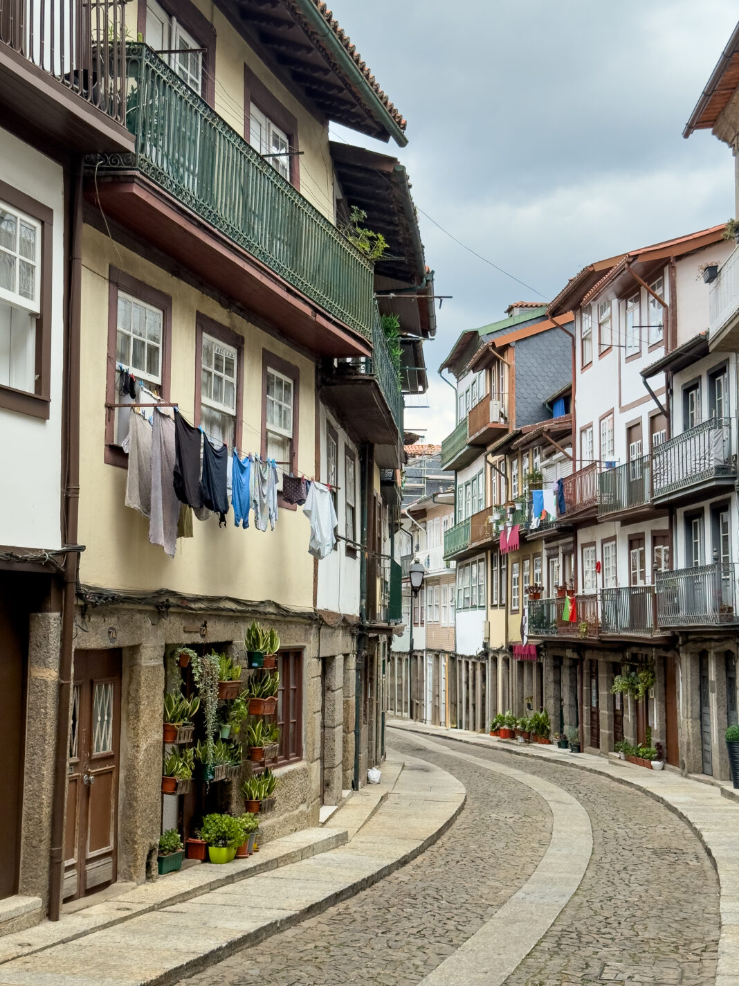 Foto: Gasse in Guimarães. Blick eine Gasse entlang, die eine Linkskurve nimmt. Links und rechts stehen dicht an dicht alte Häuschen. die Wäsche, Fahnen udn Balkone an der Fassade haben.